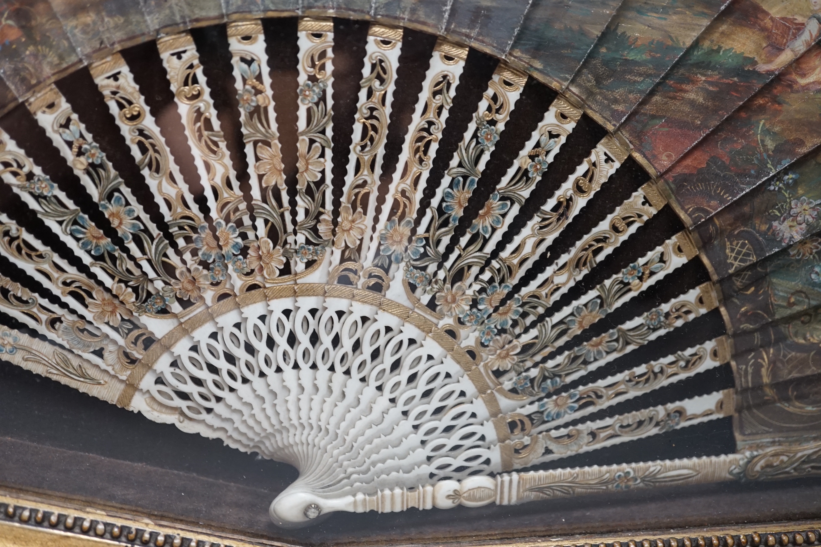 A 19th century French bone fan with pierced decoration, hand painted with figures wearing 18th century dress before a landscape, framed, the frame 62cm wide